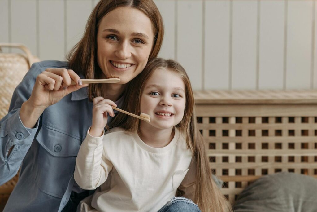 Mother and daughter bond while smiling and holding wooden toothbrushes indoors.
