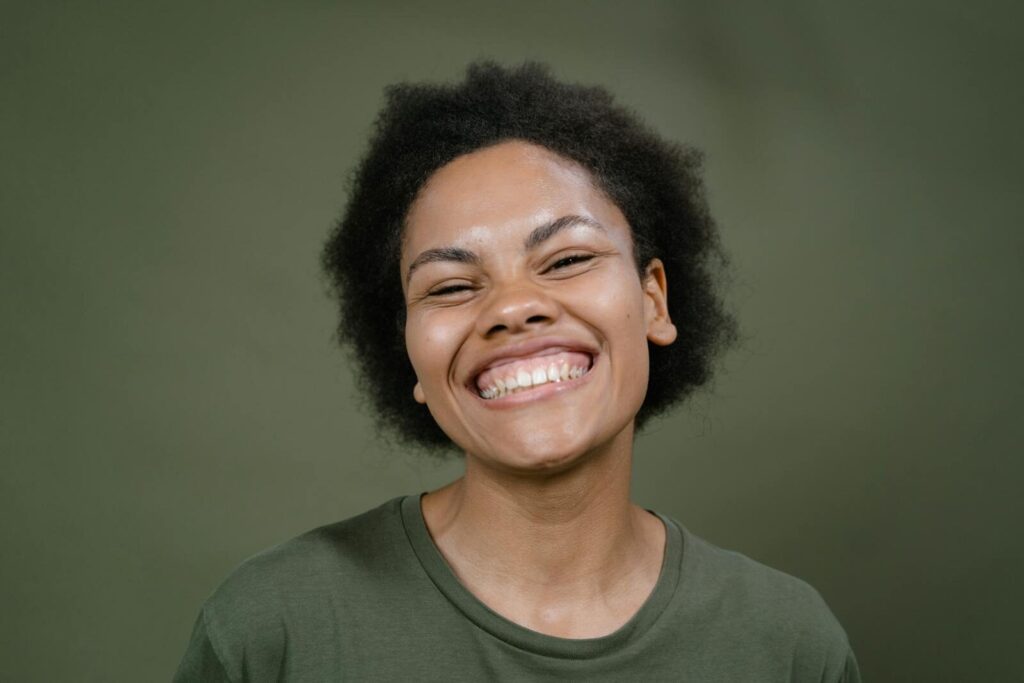 A cheerful woman with afro hair smiles broadly against a green background, exuding happiness.